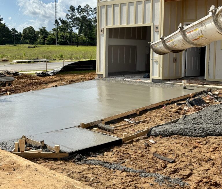Partially built two-story house with board-and-batten siding and a newly poured concrete slab driveway, with a concrete delivery chute extending into wooden formwork on a dirt construction site.