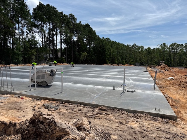 Commercial construction site: freshly poured concrete slab foundation being finished with a power trowel, utility pipes protruding, and workers in high-visibility gear under a blue sky