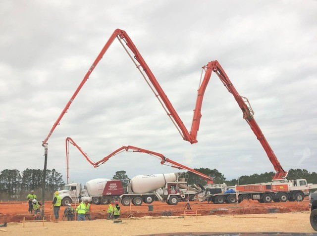 Government construction project site with workers operating large concrete pump trucks from American Concrete Supply, pouring concrete from mixers into the foundation area